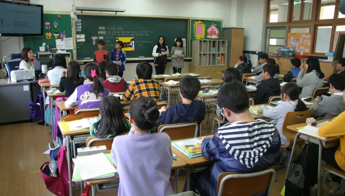 Sala de aula cheia vista do fundo, com alunos voltados para a lousa